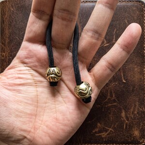 May include: Two brass beads with intricate engraved patterns threaded onto a black braided cord. The metallic beads feature a rounded, textured design with visible grooves and details, securely attached to the ends of the black rope material.