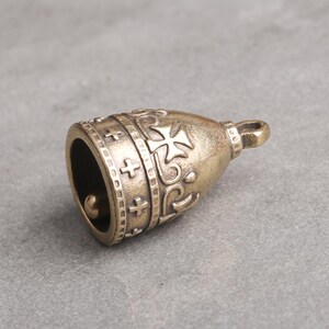 May include: A brass-toned metal bell charm featuring a rounded shape with embossed cross symbols and decorative scrollwork patterns. The bell has a small loop at the top for attachment and a clapper visible inside the hollow opening at the base.