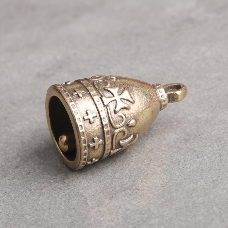 May include: A brass-toned metal bell charm featuring a rounded shape with embossed cross symbols and decorative scrollwork patterns. The bell has a small loop at the top for attachment and a clapper visible inside the hollow opening at the base.
