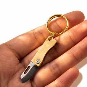 Op de afbeelding: Een miniatuur opvouwbaar zakmes als sleutelhanger met een goudkleurig metalen handvat, een enkele zilverkleurige schroef en een klein, scherp stalen lemmet met een gleuf, bevestigd aan een ronde, goudkleurige metalen sleutelring.
