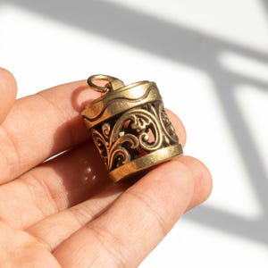 May include: A cylindrical brass pendant featuring an intricate openwork filigree design with swirling patterns. The top of the pendant has a smooth band with a wavy engraved line and a small circular jump ring attached for hanging on a chain.