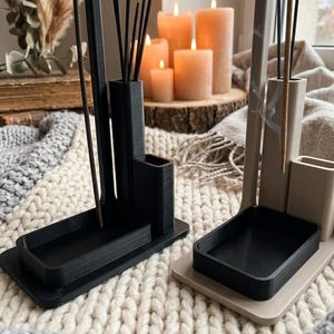 Peut inclure: Deux porte-encens, un noir et un beige, chacun doté d'une base rectangulaire, d'un tube vertical haut pour les bâtonnets d'encens et d'un compartiment rectangulaire plus court. Les supports présentent une texture en couches visible.
