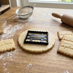 Peut inclure: Un emporte-pièce rectangulaire en plastique noir avec un bord festonné et le nom "justin" en écriture cursive avec de petits cœurs de chaque côté. L'emporte-pièce est posé sur une pâte crue ronde, entouré de biscuits cuits avec la même empreinte.