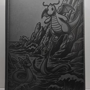 May include: Gray binder featuring a detailed illustration of a dragon-like creature soaring above the ocean, with two similar creatures in the water. The artwork is etched into the binder's surface, creating a textured, monochromatic design.