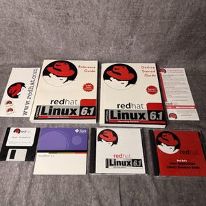 May include: A collection of Red Hat Linux 6.1 software materials including a Reference Guide, Getting Started Guide, a boot diskette, a StarOffice 5.1 disc, a Linux 6.1 disc, and a Library Resource Guide, all featuring the red fedora logo.