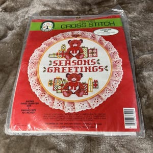 May include: A Colortex stamped cross stitch kit in a plastic package. The kit features a design of two teddy bears with gifts and the text Seasons Greetings. The package indicates a finished size of 10.5 inches and includes an 8 inch wooden hoop.