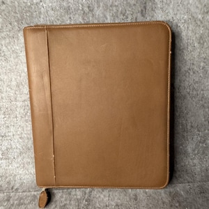 May include: A rectangular brown leather portfolio case with a smooth finish and a vertical stitched seam along the left side. The case features a zipper closure that runs along the top and right edges, with a small matching brown leather zipper pull.