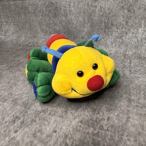 May include: A plush caterpillar toy featuring a bright yellow head with a round red nose and two blue antennae. The body is segmented with green, blue, and red fabric sections, and it has multiple soft green legs extending from the sides.