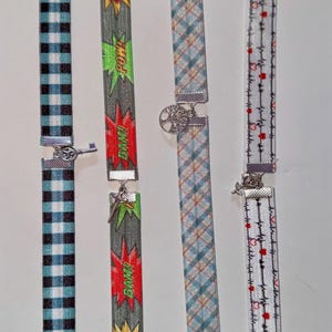 Puede incluir: Cuatro bandas elásticas de tela con cierres de metal plateado. La primera tiene un patrón de cuadros vichy azul y negro con un dije de llave. La segunda presenta texto estilo cómic "POW" y "BAM". La tercera tiene un patrón de cuadros azul con un dije de árbol. La cuarta muestra una línea de latido negra con corazones rojos.