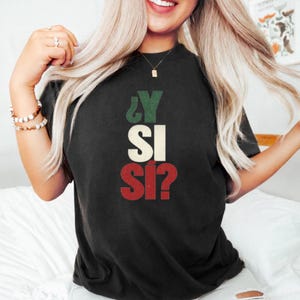 Può includere: Una t-shirt nera a maniche corte con il testo ¿Y SI SÍ? stampato in stile vintage. La lettera Y è verde, le lettere SI sono bianche e le lettere SÍ sono rosse, disposte verticalmente al centro della zona del petto.