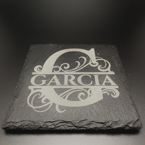 Pode incluir: Um porta-copos quadrado de ard&oacute;sia preta com um design gravado a laser. O centro apresenta uma letra G mai&uacute;scula grande com ornamentos decorativos. O nome GARCIA est&aacute; gravado numa linha horizontal atrav&eacute;s do centro da letra G.