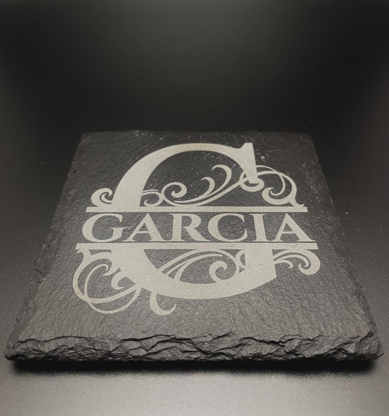 Pode incluir: Um porta-copos quadrado de ard&oacute;sia preta com um design gravado a laser. O centro apresenta uma letra G mai&uacute;scula grande com ornamentos decorativos. O nome GARCIA est&aacute; gravado numa linha horizontal atrav&eacute;s do centro da letra G.