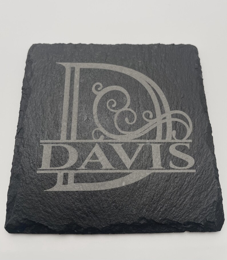 Pode incluir: Um porta-copos quadrado de ard&oacute;sia preta com uma letra D grande gravada com detalhes decorativos em volutas. O nome DAVIS est&aacute; gravado numa fonte serifada em negrito e mai&uacute;sculas no centro da letra D, criando um design personalizado.