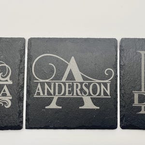 Pode incluir: Tr&ecirc;s porta-copos quadrados de ard&oacute;sia preta com desenhos gravados a laser. Cada porta-copos exibe uma grande letra inicial decorativa com uma linha horizontal no centro contendo os nomes GARCIA, ANDERSON e DAVIS.