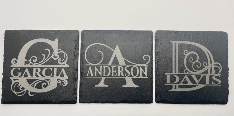 Pode incluir: Tr&ecirc;s porta-copos quadrados de ard&oacute;sia preta com desenhos gravados a laser. Cada porta-copos exibe uma grande letra inicial decorativa com uma linha horizontal no centro contendo os nomes GARCIA, ANDERSON e DAVIS.