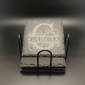 Pode incluir: Uma pilha de porta-copos quadrados de ard&oacute;sia preta com bordas naturais irregulares num suporte de arame met&aacute;lico preto. O porta-copos superior apresenta uma letra G grande gravada com o nome GARCIA centrado numa fonte com serifa.