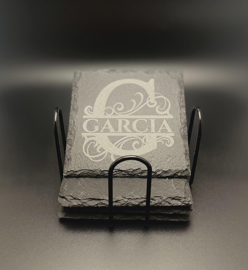 Pode incluir: Uma pilha de porta-copos quadrados de ard&oacute;sia preta com bordas naturais irregulares num suporte de arame met&aacute;lico preto. O porta-copos superior apresenta uma letra G grande gravada com o nome GARCIA centrado numa fonte com serifa.
