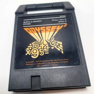 Könnte beinhalten: Schwarze Odyssey 2 Videospielkassette mit dem Text "MATH-A-MAGIC! ECHO!" und orangefarbenen und gelben Grafiken. Die Kassette hat einen Griff und enthält Text wie "Press 1 thru 3" und "Press 4". Copyright 1978 N.A.P. Consumer Electronics Corp.