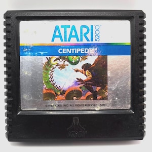 Peut inclure: Cartouche de jeu vidéo Atari 5200 Centipede noire. L'étiquette présente le logo Atari, le titre du jeu et une illustration colorée d'un mille-pattes et d'une figurine. La cartouche est dans un boîtier en plastique noir avec un design strié.