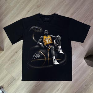 Può includere: Una t-shirt nera a maniche corte con una stampa grafica di un giocatore di basket con una maglia gialla e viola dei Lakers con il numero 24, seduto su un pallone da basket avvolto da un grande serpente nero, con una firma bianca sul davanti.