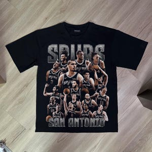 Könnte beinhalten: Ein schwarzes Kurzarm-T-Shirt mit einer Collage von San Antonio Spurs Basketballspielern in ihren Teamtrikots. Die Grafik zeigt den Schriftzug "SPURS" oben und "SAN ANTONIO" unten in einer abgenutzten Schriftart im Metallic-Look.