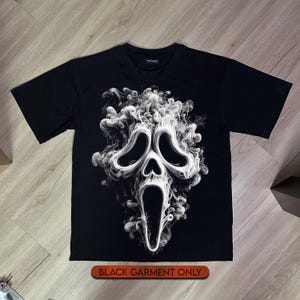 May include: A black short-sleeved t-shirt featuring a large, white, swirling smoke graphic in the shape of a ghost mask with hollow eyes and an open mouth. The text "BLACK GARMENT ONLY" is printed in a small orange rectangular box below the shirt.