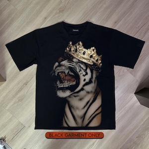 Puede incluir: Una camiseta negra de manga corta con un estampado gráfico de un tigre blanco rugiendo que lleva una corona dorada con detalles negros. El tigre muestra los dientes, que parecen estar incrustados con cristales plateados. El texto BLACK GARMENT ONLY es visible.