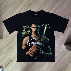 Könnte beinhalten: Ein schwarzes Kurzarm-T-Shirt mit einem Grafikdruck eines Basketballspielers im Spurs-Trikot, der einen Basketball hält. Das Design zeigt den Schriftzug The Alien, ein grün leuchtendes UFO und eine teilweise Alien-Transformation am Körper.