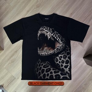 Könnte beinhalten: Ein schwarzes T-Shirt mit kurzen Ärmeln und einem großen Grafikdruck einer Schlange mit offenem Maul. Die Schlange hat ein geflecktes Muster und ihre Zähne sind mit einer schimmernden, diamantartigen Textur dargestellt. Text: BLACK GARMENT ONLY.