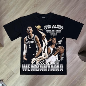 Könnte beinhalten: Ein schwarzes Kurzarm-T-Shirt mit einem Grafikdruck des Basketballspielers Victor Wembanyama im Trikot der San Antonio Spurs. Das Design enthält den Text THE ALIEN, SAN ANTONIO SPURS und WEMBANYAMA sowie einen Planeten und Sterne.
