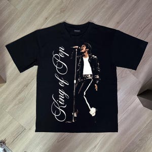 May include: A black short-sleeve t-shirt featuring a graphic print of a performer singing into a microphone. The text "King of Pop" is printed in a large white cursive font vertically along the left side of the chest area of the garment.
