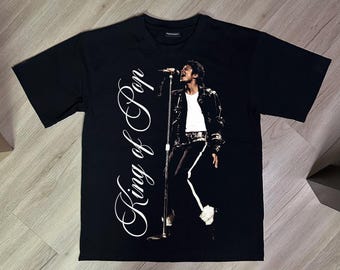 Peut inclure: Un t-shirt noir à manches courtes avec une impression graphique représentant un artiste chantant dans un microphone. Le texte "King of Pop" est imprimé en grande police cursive blanche verticalement sur le côté gauche de la poitrine.