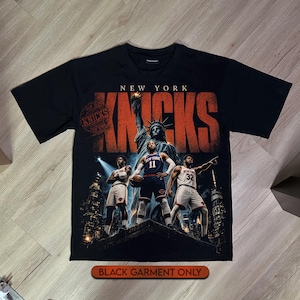 Può includere: Una t-shirt nera con una stampa grafica dei New York Knicks e il testo NEW YORK KNICKS. Il design include la Statua della Libertà, tre giocatori di basket in divisa e uno sfondo con lo skyline della città e un logo a timbro circolare.