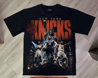 May include: A black t-shirt featuring a graphic print of the New York Knicks with the text NEW YORK KNICKS. The design includes the Statue of Liberty, three basketball players in uniform, and a city skyline background with a circular stamp logo.
