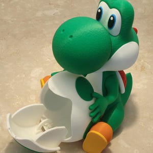 Op de afbeelding: Een groene Yoshi-figuur met een witte eierschaal. De figuur heeft grote blauwe ogen, een rood zadel en oranje schoenen. De eierschaal is open en onthult de binnenkant. Het beeldje is een personage uit de Super Mario-videogame.