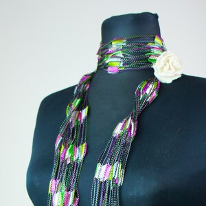May include: A multi-strand scarf with a green, pink, and white pattern. The scarf is made of a metallic material and has a delicate, flowing texture. It is displayed on a black mannequin.