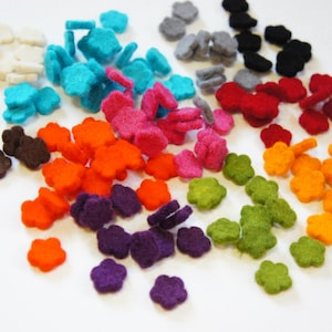 Op de afbeelding: Assortiment van viltbloemvormen in verschillende kleuren, waaronder wit, turquoise, roze, oranje, paars, groen, rood en bruin. De viltbloemen zijn verspreid over een wit oppervlak.