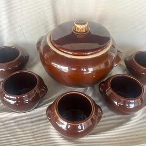 May include: A set of dark brown glazed ceramic kitchenware consisting of one large lidded tureen with two side handles and four matching smaller bowls, each featuring two small side handles and a smooth, glossy finish throughout the entire collection.