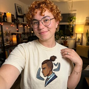 K&ouml;nnte beinhalten: Cremefarbenes T-Shirt mit einer Cartoon-Illustration eines Charakters mit einem Dutt-Haarschnitt und blauen Akzenten. Der Charakter ist im Profil mit einem strengen Ausdruck dargestellt. Das T-Shirt wird von einer Person mit lockigem rotem Haar und Brille getragen.