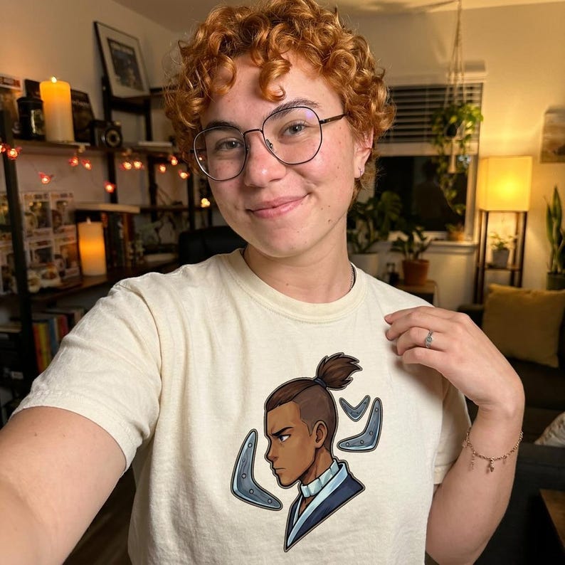 K&ouml;nnte beinhalten: Cremefarbenes T-Shirt mit einer Cartoon-Illustration eines Charakters mit einem Dutt-Haarschnitt und blauen Akzenten. Der Charakter ist im Profil mit einem strengen Ausdruck dargestellt. Das T-Shirt wird von einer Person mit lockigem rotem Haar und Brille getragen.