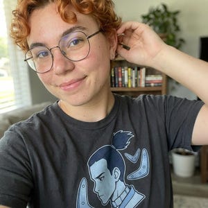 Pu&ograve; includere: T-shirt grigio scuro con un personaggio dei cartoni animati blu e boomerang blu. Il personaggio &egrave; rivolto verso sinistra. La persona nella foto indossa occhiali rotondi con montatura nera.