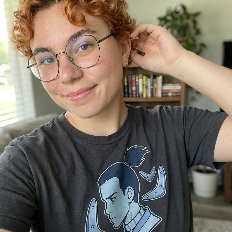 Pu&ograve; includere: T-shirt grigio scuro con un personaggio dei cartoni animati blu e boomerang blu. Il personaggio &egrave; rivolto verso sinistra. La persona nella foto indossa occhiali rotondi con montatura nera.