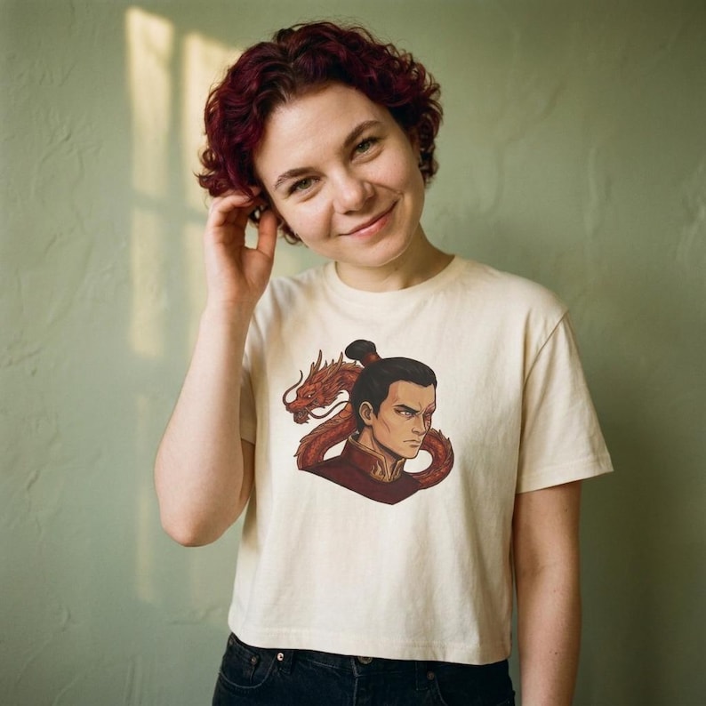 May include: A cream-colored short-sleeved t-shirt featuring a graphic illustration of a stern-faced character with a scar over one eye, wearing a dark red high-collared garment, positioned in front of a coiled red dragon with detailed scales.