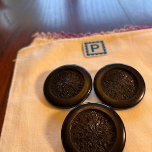 Può includere: Tre bottoni rotondi neri con un centro strutturato, disposti su un tessuto color crema. È visibile anche un piccolo quadrato bordato di blu con la lettera "P". I bottoni hanno una finitura lucida.