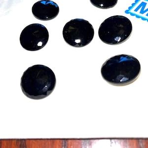 May include: Seven faceted, oval-shaped black gemstones are arranged on a white surface. The gems have a glossy finish and reflect light, creating highlights. The arrangement suggests they could be loose stones for jewelry making or other crafts.