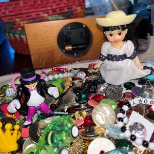 May include: A collection of assorted craft items including a small doll in a white dress and straw hat, a miniature figure in a black top hat and purple pants, a translucent green toy car, a yellow crown, and various colorful buttons and beads.