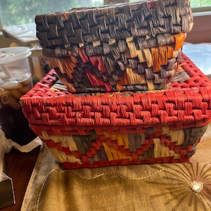 May include: Two rectangular woven baskets stacked on top of each other. The top basket features a dark brown, orange, and tan geometric pattern. The bottom basket has a vibrant red rim and a base with a multi-colored pattern of red, tan, and brown.
