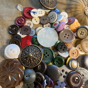 Puede incluir: Una gran colección de botones de costura surtidos en varias formas, tamaños y colores, incluyendo estilos redondos, planos y abombados. El surtido presenta materiales como plástico, metal y madera, con algunos botones que muestran patrones en relieve.