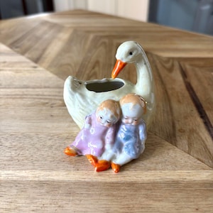 Peut inclure: Une figurine en céramique représentant un cygne crème au bec orange, avec deux petits enfants assis devant le corps du cygne. Un enfant porte une tenue rose et l'autre une tenue bleue, tous deux avec des chaussures orange, finition brillante.