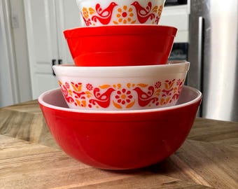 May include: A stack of four nesting mixing bowls. The top and third bowls are white with a repeating orange and red pattern of birds and flowers. The second and bottom bowls are solid bright red. The bowls are round with smooth edges.